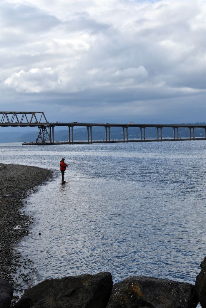 A solitary fisherman casts his line by the sea with a bridge in the background under a cloudy sky.